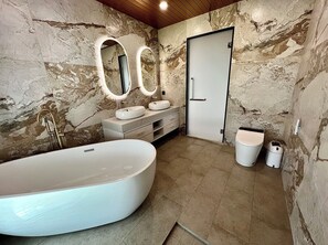 Ein Badezimmer mit freistehender Badewanne, zwei Waschbecken mit runden Spiegeln, einer Tür und einem Mülleimer.