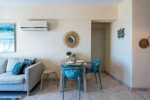 Uma sala de estar com sofá, mesa de jantar com cadeiras, ar-condicionado fixado na parede, um espelho decorativo e um letreiro motivacional.