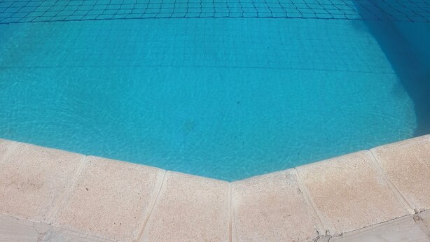 Una piscina rectangular con una barrera de seguridad de malla, rodeada por un suelo de baldosas y una cerca blanca.