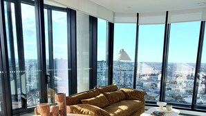Uma sala de estar moderna com uma mesa redonda de madeira, um sofá marrom e vista para um horizonte urbano através de grandes janelas.