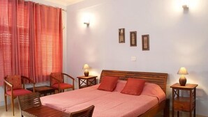 A bedroom with a bed, two chairs, a table, and two bedside tables with lamps. - Ayurveda Bhavan (Rishikesh)