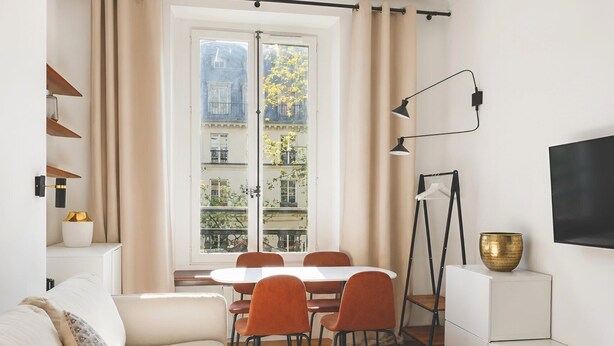 A modern living room with a sofa, dining table, chairs, a coffee table, a TV, and a window with a view of buildings and trees. - 345-Tronchet 1G - Stunning Apartment in Paris 8 (Paris)