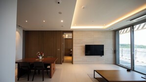 A modern living room with a large flat-screen TV mounted on the wall, a dining area with a wooden table and chairs, and a large window offering a view of the outdoors. - Haeundae Beach Restmons (Busan)