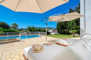 Ein Poolbereich mit Liegestuhl, Sonnenschirm und Blick auf ein Schwimmbecken und Grünflächen.