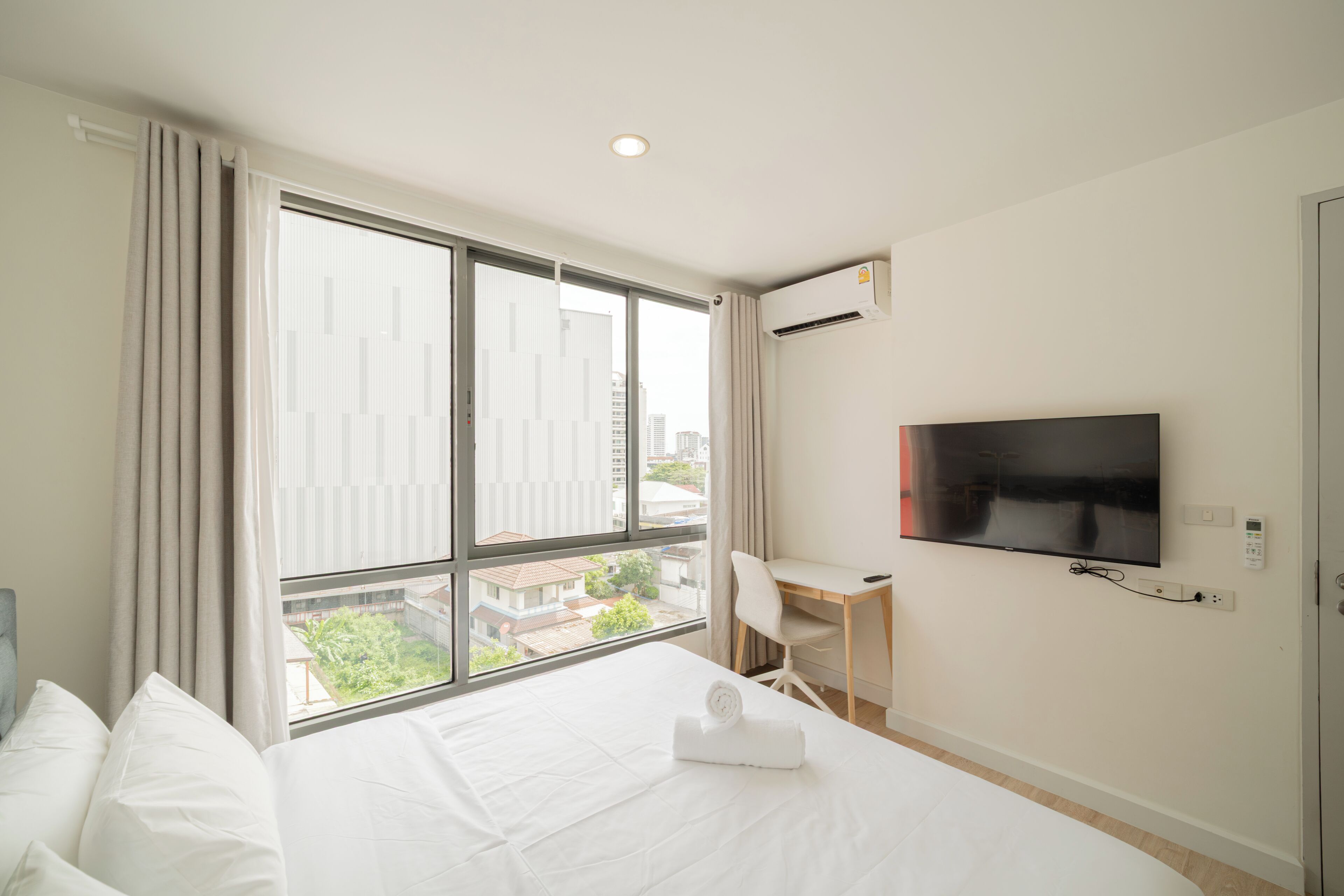 Une chambre d’hôtel dotée d’une grande fenêtre, d’un lit avec du linge de maison blanc, d’un bureau avec une chaise, d’un téléviseur à écran plat fixé au mur et d’un appareil de climatisation.