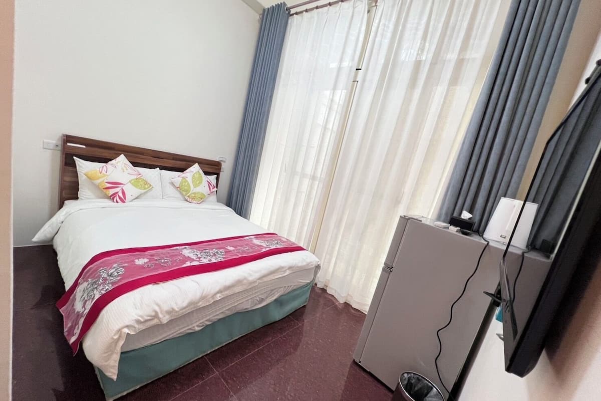 Una habitación de hotel con cama, cortinas, un escritorio con computadora y un cubo de basura.