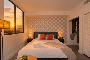A hotel room with a large bed, two bedside tables, a view of the sunset, and a patterned wallpaper.