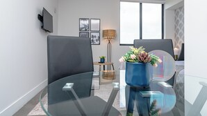 Ein modernes Wohnzimmer mit zwei grauen Sesseln, einem Glastisch, einer blauen Vase mit Blumen, einer Stehlampe und einem an der Wand befestigten Flachbildfernseher.