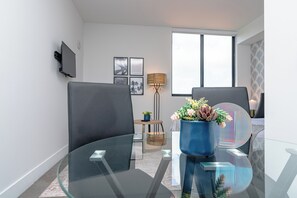 Ein modernes Wohnzimmer mit zwei grauen Sesseln, einem Glastisch, einer blauen Vase mit Blumen, einer Stehlampe und einem an der Wand befestigten Flachbildfernseher.