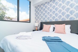 Ein Hotelzimmer mit einem Bett, Kissen, einem Nachttisch, einer Lampe und einem Fenster mit Blick auf den Himmel und ein Gebäude.