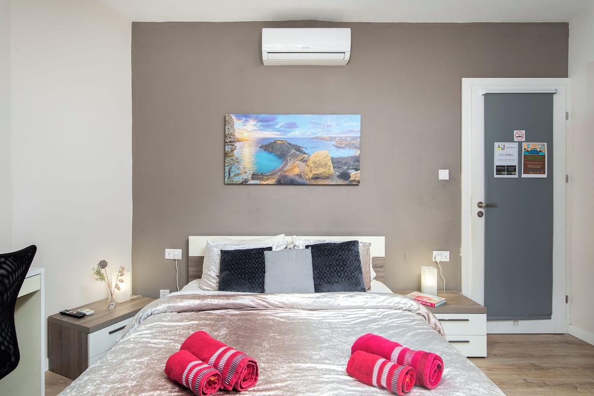 Une chambre d’hôtel comprenant un lit, une table de chevet, une commode, un climatiseur fixé au mur, une porte avec une pancarte et un tableau accroché au mur.