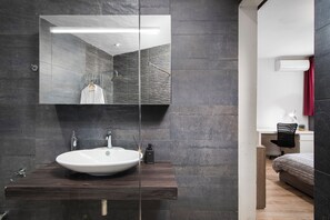 A modern bathroom with a large mirror, dark stone tiles, a white sink, and a wooden vanity. - Luxury Msida Circle Suites (Mgarr)