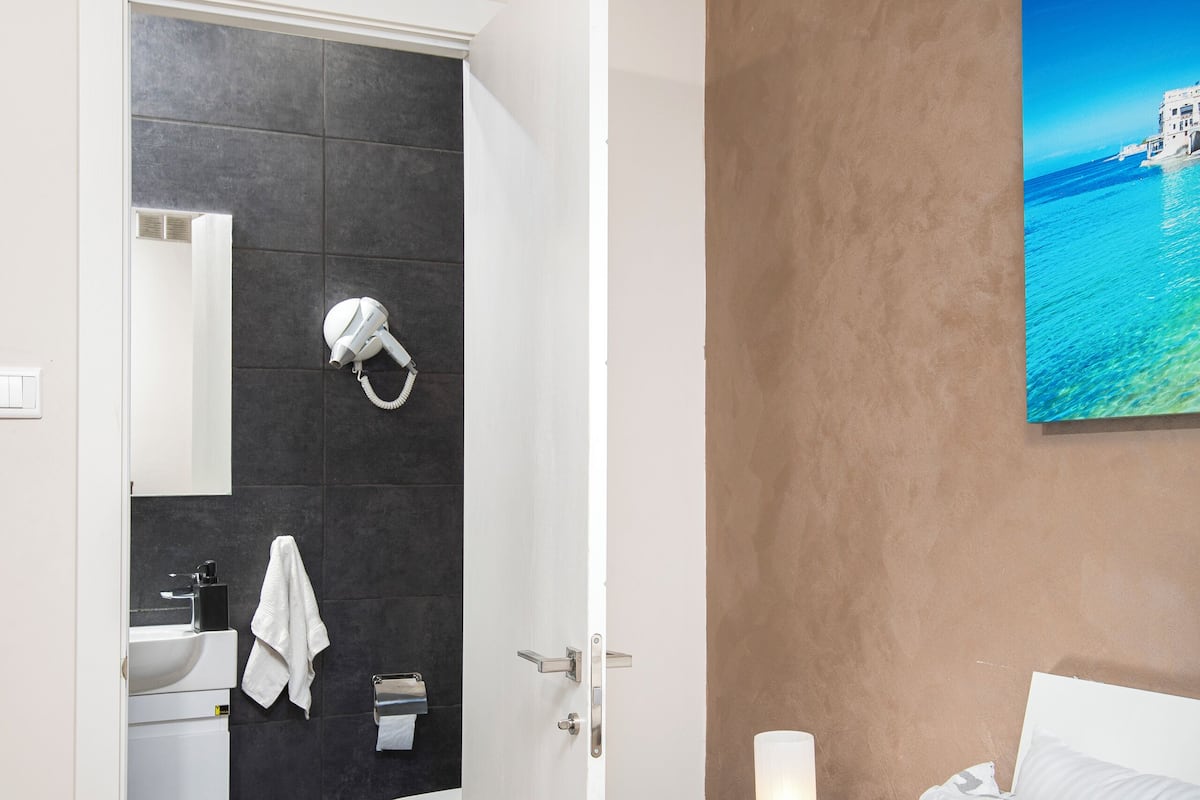 Une chambre d’hôtel moderne, équipée d’un lit, d’une table de chevet, d’une salle de bain avec un lavabo et des toilettes, et ornée d’un tableau au mur.