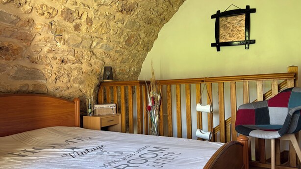 Ein Schlafzimmer mit Steinwand, einem hölzernen Bett, einem Nachttisch mit Lampe und einem Sessel mit gesteckter Decke.