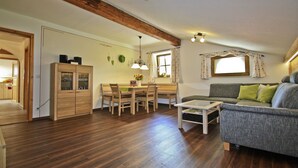 Amplia sala de estar con piso de madera, un sofá gris, una mesa de comedor con sillas y una zona de cocina con armario de madera.