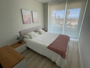 A bedroom with a bed, bedside tables, a nightstand, and a window with a view of a parking lot and palm trees. - Swing &Sun Free Parking by StayCare (Chipiona)