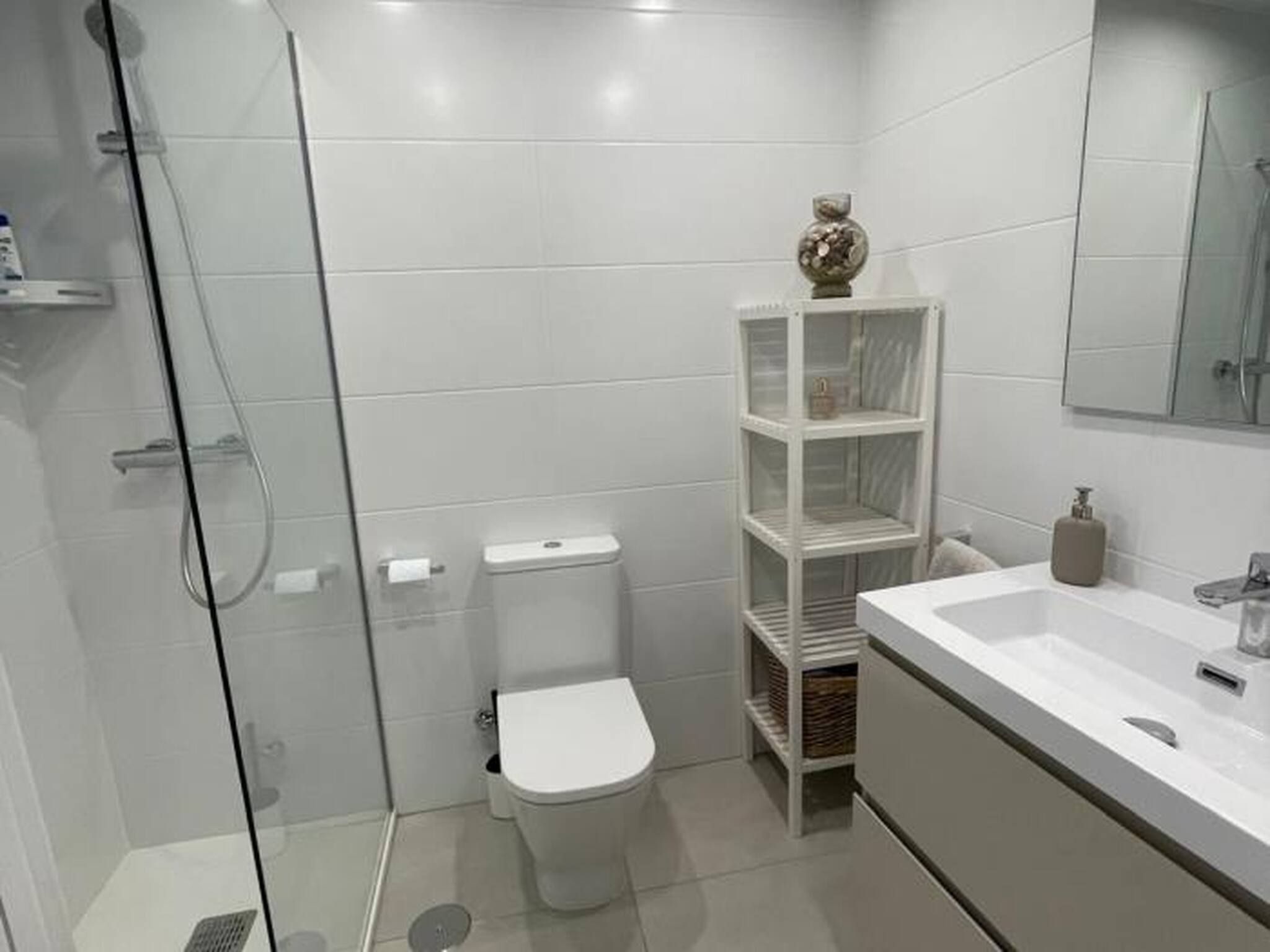 A bathroom with a glass-enclosed shower, a toilet, a white sink with a mirror, and a white shelving unit with a decorative item on top.