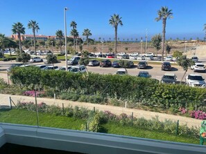 Ein Parkplatz mit mehreren Autos, ein umzäunter Gartenbereich mit Sträuchern und Blumen und ein freier Blick auf das Meer im Hintergrund.