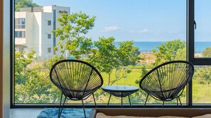 Une pièce avec deux chaises au design minimaliste en fil de fer noir, une petite table ronde et une vue sur un immeuble et de la verdure, le tout visible à travers une grande fenêtre.