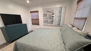Ein Schlafzimmer mit einem Bett, einem Schminktisch, einem Fernseher und zwei Fenstern mit Jalousien.