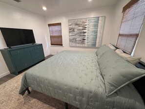 Ein Schlafzimmer mit einem Bett, einem Schminktisch, einem Fernseher und zwei Fenstern mit Jalousien.