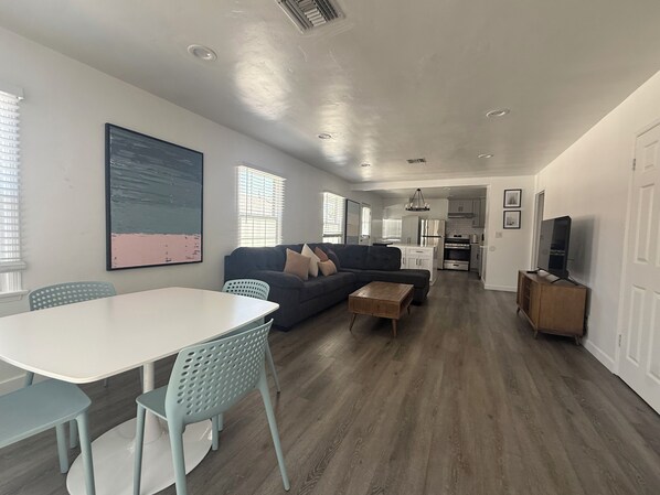 Een moderne woonkamer met een bank, een eettafel en stoelen, een keuken op de achtergrond en een televisie op een kast.