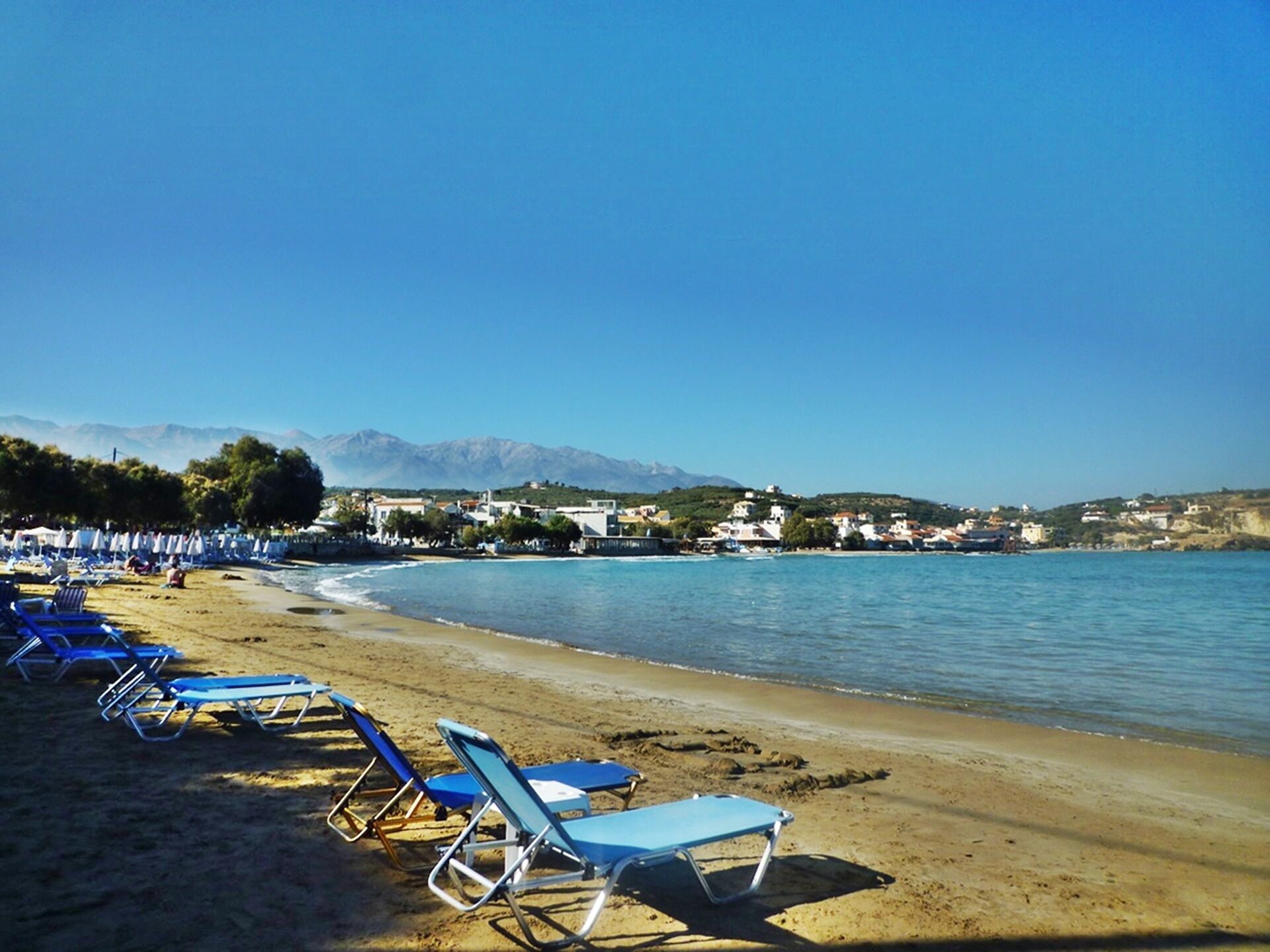 Une plage bordée de rangées de chaises longues bleues, un rivage sablonneux et des bâtiments en arrière-plan.