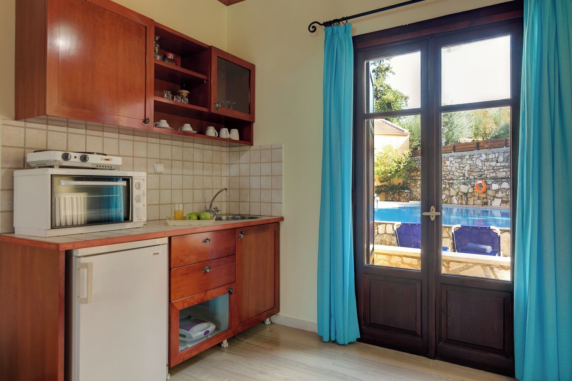 Una cocina con armarios de madera, microondas, refrigerador y ventana con cortinas.