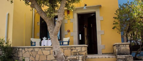 Um edifício amarelo com degraus de pedra que levam a uma porta, uma árvore em primeiro plano e uma mesa com cadeiras do lado de fora.