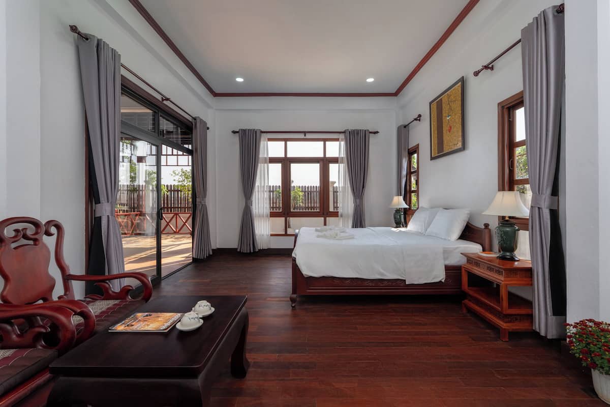 A hotel room with a bed, a wooden table, a chair, a window with curtains, and a view of the outside.