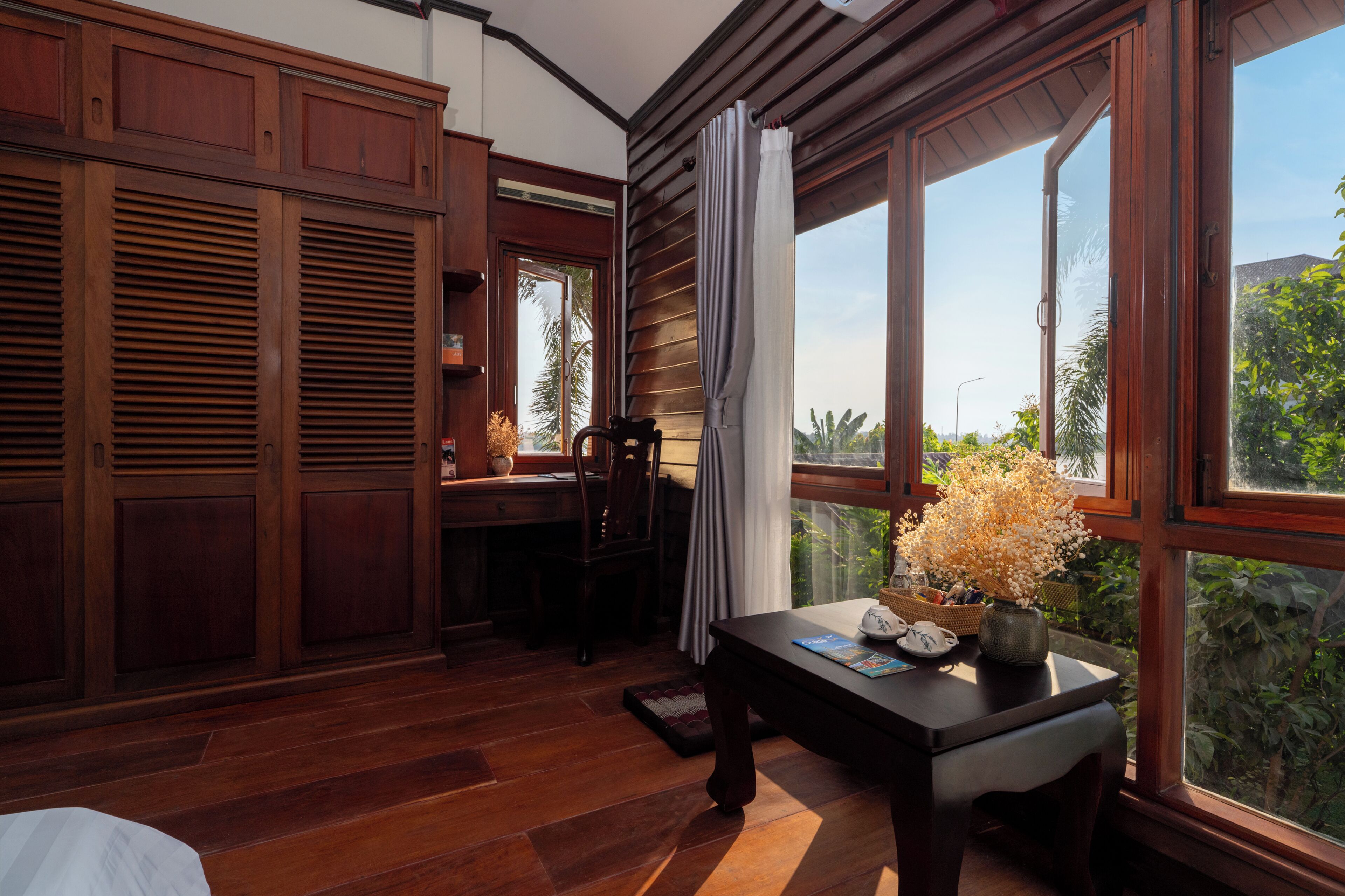 A room with wooden flooring, a desk with a vase of flowers, a chair, and large windows with curtains.