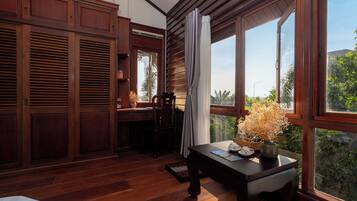A room with wooden flooring, a desk with a vase of flowers, a chair, and large windows with curtains.