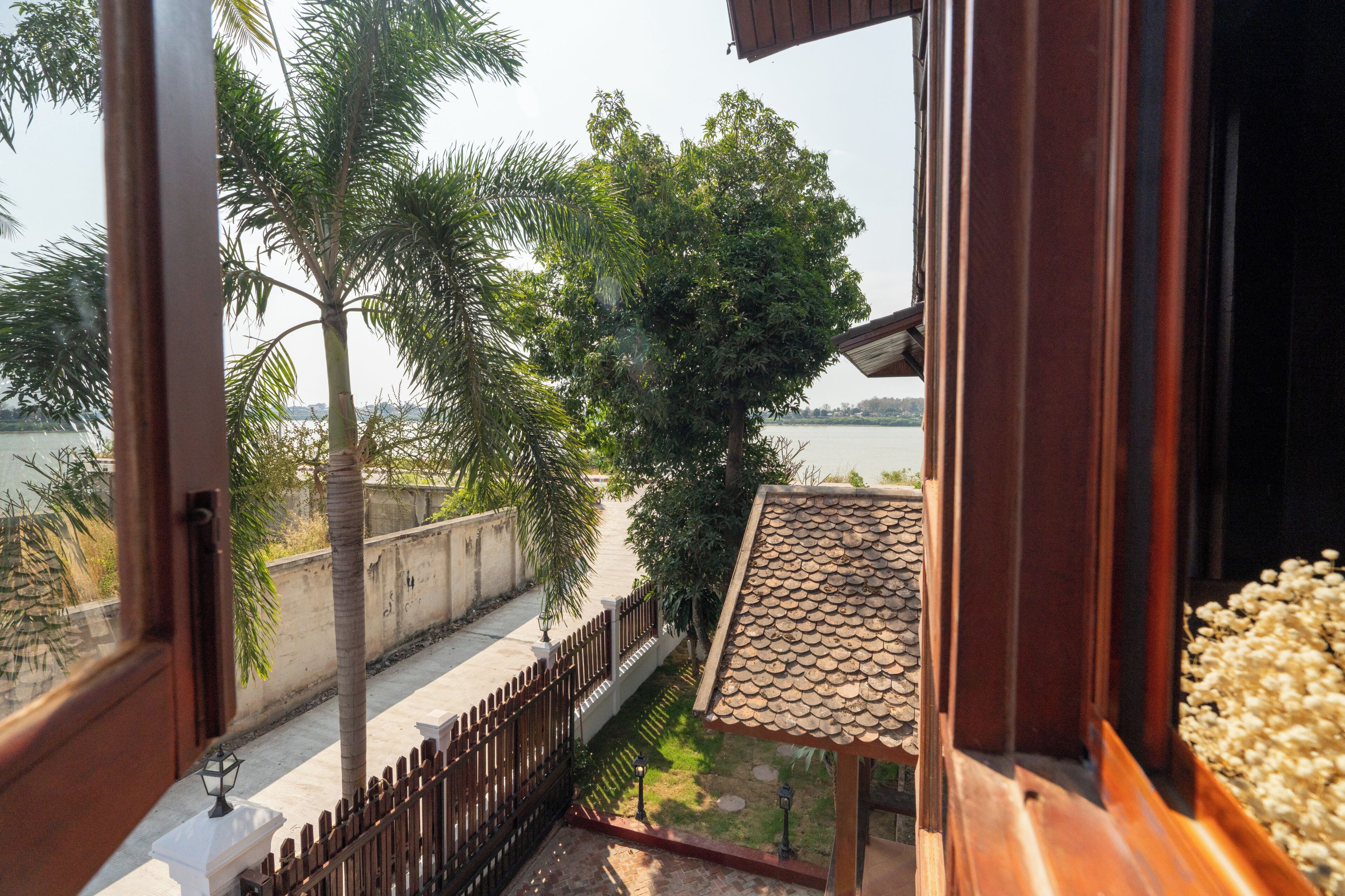 A view from a room with a window overlooking a pathway, palm trees, and a body of water.
