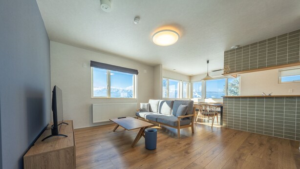 A modern living room with a sofa, coffee table, and TV stand. There are large windows and a view of the outdoors. - Furano Entire House｜Ski & Lavender｜Mountain View｜Groups up to 10 ｜ 3BR (Furano City)