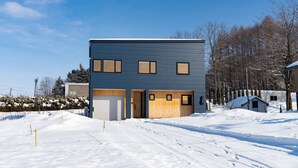 A modern two-story building with a garage, a covered driveway, and a snow-covered landscape. - Furano Entire House｜Ski & Lavender｜Mountain View｜Groups up to 10 ｜ 3BR (Furano City)