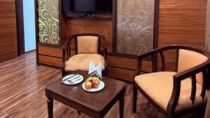 A room with a wooden floor, two wooden chairs, a small table with fruit, and a flat-screen TV mounted on the wall. - The Amberly Gardens (Sanwer)