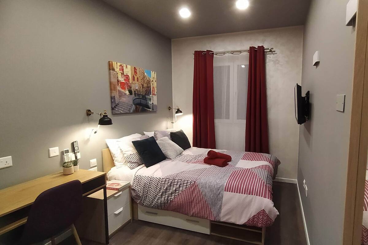 A bedroom with a bed, a desk, a chair, a painting, and a window with curtains.