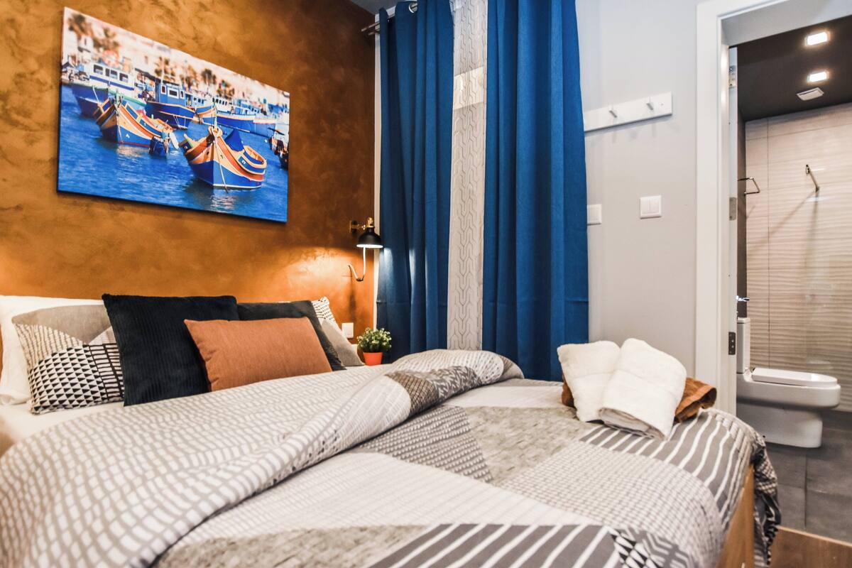 A hotel room with a bed, a nightstand, a painting of boats, a shower, and a toilet.