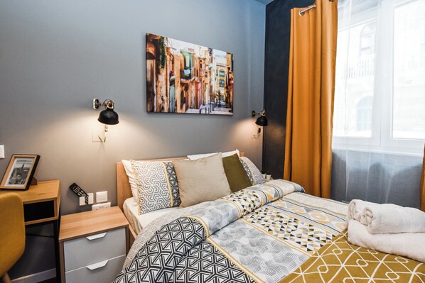 A hotel room with a bed, bedside lamps, a desk, a chair, a framed picture, and a window with curtains. - Cosy Albert's Seaside Suites - Sliema (Gżira)
