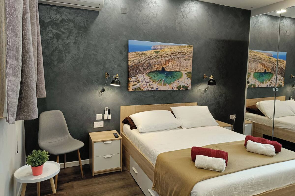 A bedroom with a bed, bedside tables, a chair, a small table with a plant, and a wall-mounted air conditioner.