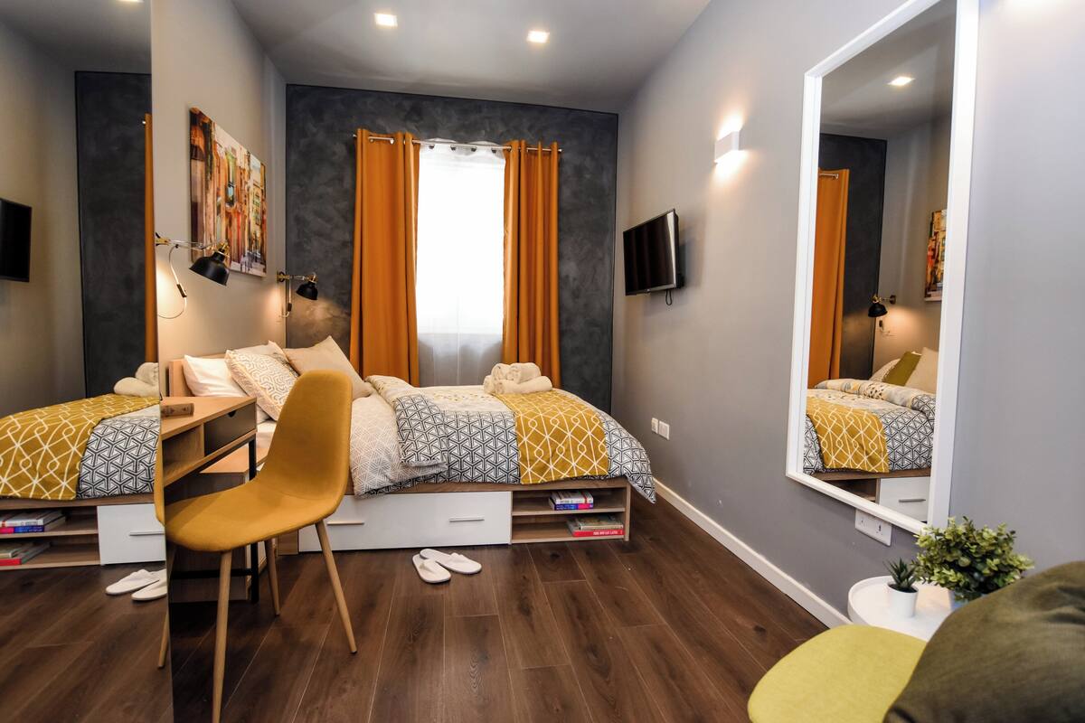 A bedroom with a bed, a desk, a chair, a mirror, and a small plant.