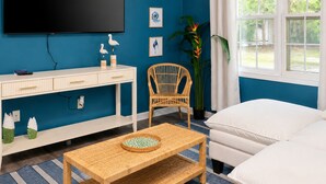 Ein Zimmer mit Fernseher, Schreibtisch, Korbsessel, Bett und Fenster mit Vorhängen.