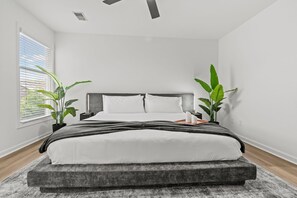 Ein modernes Schlafzimmer mit einem Bett, zwei Topfpflanzen, einem Deckenventilator und einem Fenster mit Jalousien.