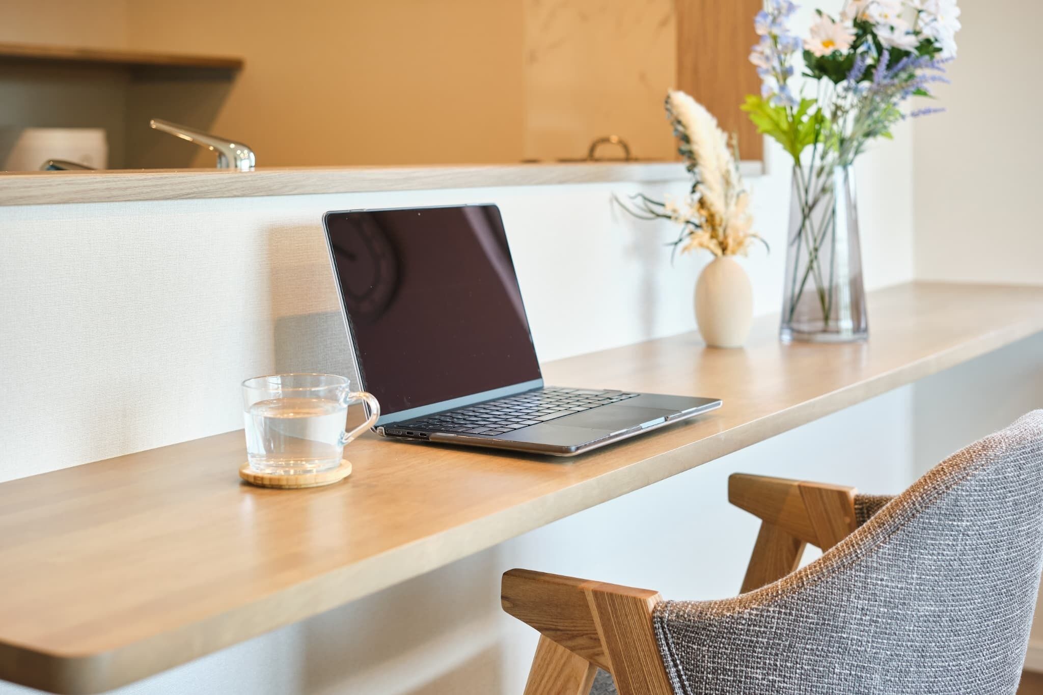 A laptop on a wooden desk with a glass of water, a vase with flowers, and a chair.