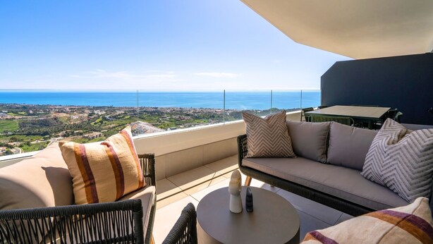 Een balkon met een bank, stoelen en een tafel, met uitzicht op een kustlandschap.