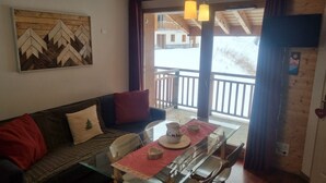 A cozy living area with a sofa, a dining table with chairs, a wooden wall decoration, and a balcony with a railing. - Apartment in Valfrejus Near Gondola (Modane)