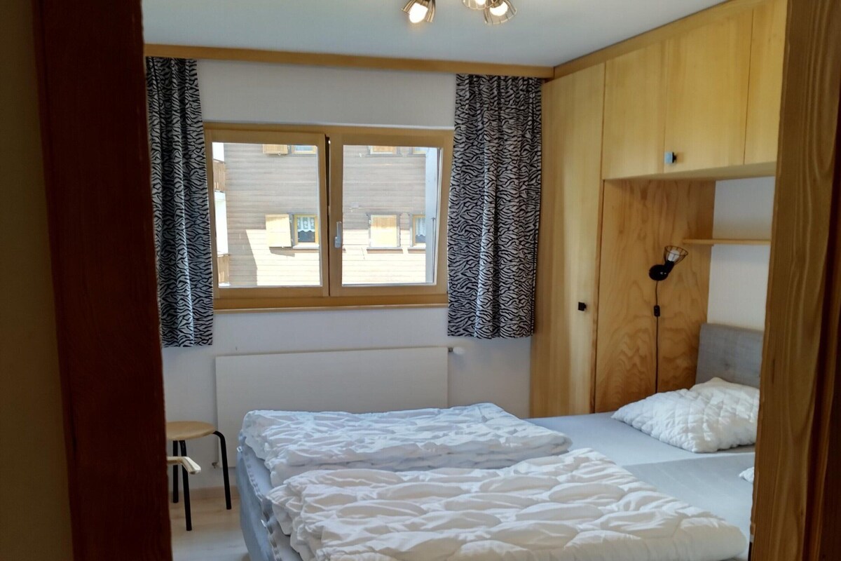 A bedroom with two beds, a window with curtains, a radiator, and a small table.
