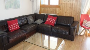 A brown leather sectional sofa with red and gray patterned cushions, a glass coffee table, wooden flooring, a wooden paneled wall, and a red chair with a patterned cushion. - Cozy Apartment for Groups (Sion)