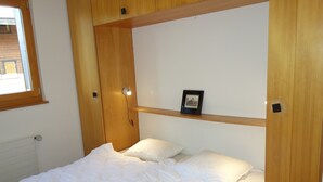 A single bed with a white duvet, wooden headboard, and a small shelf with a picture frame. - Cozy Apartment for Groups (Sion)