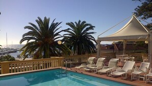 Ein Swimmingpool mit Liegestühlen und Sonnensegel, umgeben von Palmen und Blick auf den Hafen.